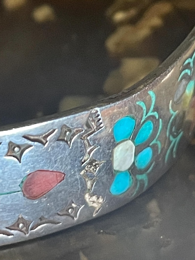 May include: Close-up of a silver bracelet with intricate floral and geometric designs. The bracelet features inlaid turquoise, white, and red accents. The design includes a red tulip and a stylized flower with turquoise petals.