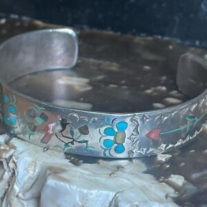 May include: A silver cuff bracelet with inlaid turquoise, red, and yellow floral designs. The open-ended bracelet features a detailed pattern of flowers and leaves, showcasing a vintage or handcrafted aesthetic. The bracelet is likely made of sterling silver.