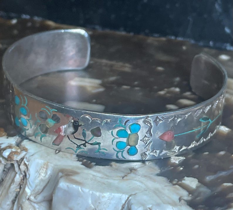 May include: A silver cuff bracelet with inlaid turquoise, red, and yellow floral designs. The open-ended bracelet features a detailed pattern of flowers and leaves, showcasing a vintage or handcrafted aesthetic. The bracelet is likely made of sterling silver.