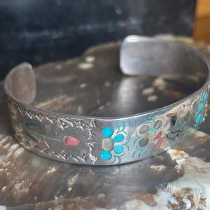 May include: Silver cuff bracelet with inlaid turquoise, coral, and mother-of-pearl floral designs. The open-ended bracelet features etched details along the edges. The bracelet is displayed on a textured surface.