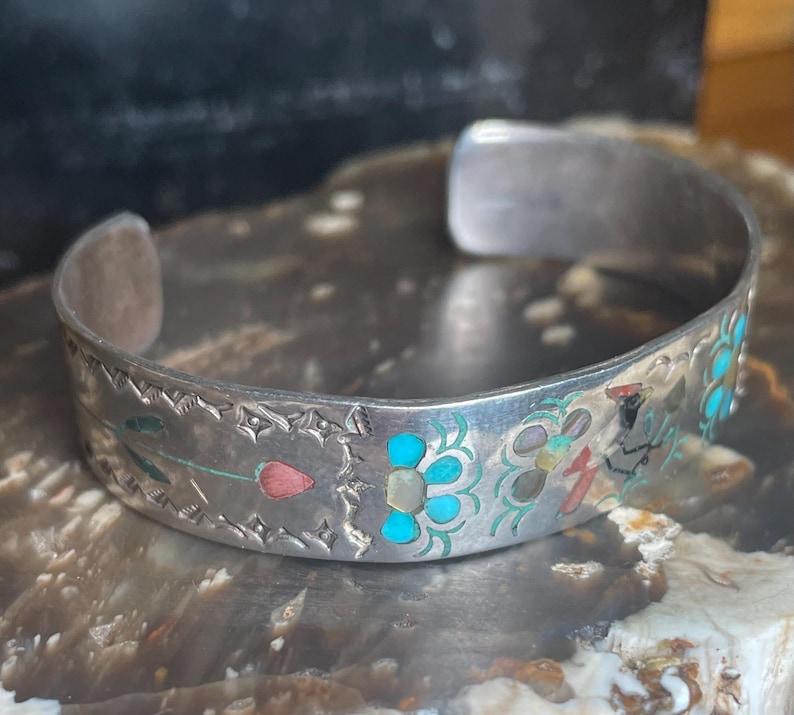 May include: Silver cuff bracelet with inlaid turquoise, coral, and mother-of-pearl floral designs. The open-ended bracelet features etched details along the edges. The bracelet is displayed on a textured surface.
