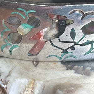 May include: Close-up of a silver bracelet with inlaid bird and floral designs. The bird features red, black, and white accents, while the flowers and leaves are turquoise. The bracelet has a polished finish and is likely vintage.