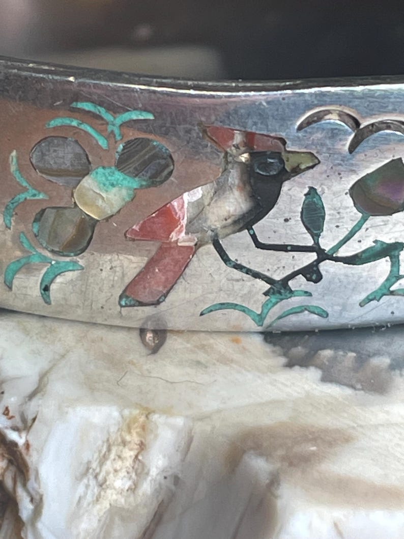 May include: Close-up of a silver bracelet with inlaid bird and floral designs. The bird features red, black, and white accents, while the flowers and leaves are turquoise. The bracelet has a polished finish and is likely vintage.