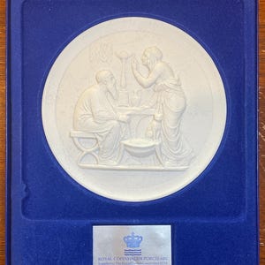 Op de afbeelding: Een wit porseleinen reliëfplaat met een klassieke scène met twee figuren en een kat, in een blauwe fluwelen doos. De plaat toont gedetailleerde figuren en objecten. De doos bevat een zilveren label met tekst en een kroonlogo.