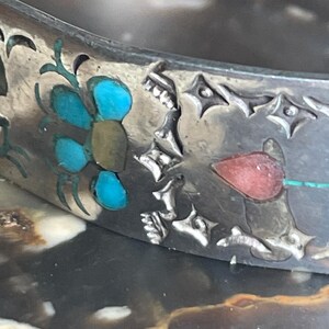 May include: Close-up of a silver bracelet with turquoise and red inlaid floral designs. The bracelet features intricate cut-out patterns and a detailed, handcrafted appearance. The design includes a flower with turquoise petals.
