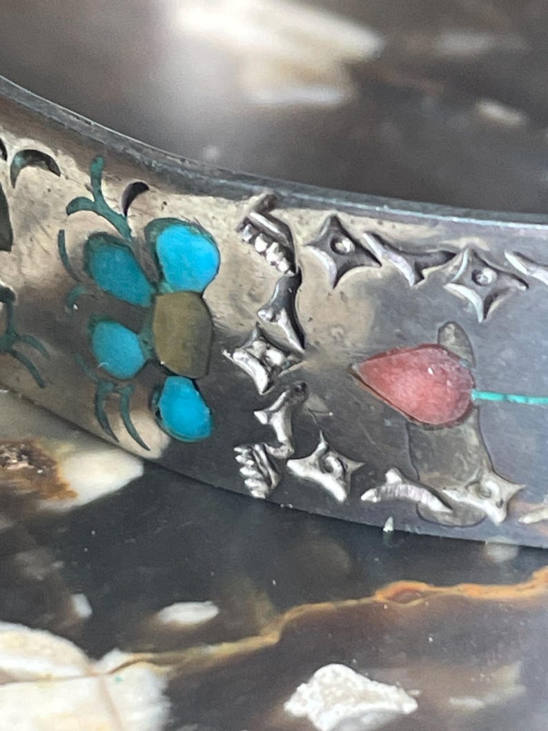 May include: Close-up of a silver bracelet with turquoise and red inlaid floral designs. The bracelet features intricate cut-out patterns and a detailed, handcrafted appearance. The design includes a flower with turquoise petals.