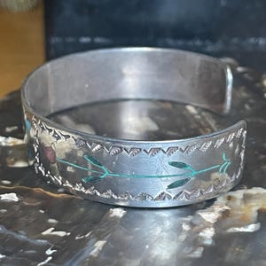 May include: Silver cuff bracelet with etched floral and geometric designs. The bracelet features turquoise and red accents, with a detailed pattern around the edges. The open design allows for easy wear.