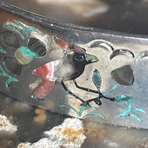 May include: Close-up of a silver bracelet with inlaid bird and floral designs. The bird features red, black, and white details, perched on a branch with turquoise accents. The bracelet has a detailed, textured surface.