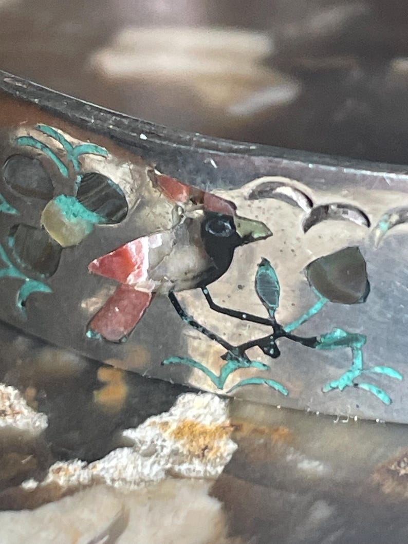 May include: Close-up of a silver bracelet with inlaid bird and floral designs. The bird features red, black, and white details, perched on a branch with turquoise accents. The bracelet has a detailed, textured surface.