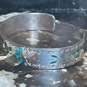 May include: Silver cuff bracelet with etched floral and geometric designs. The bracelet features turquoise, red, and green accents. The open-ended design allows for easy wear. A unique piece of jewelry.