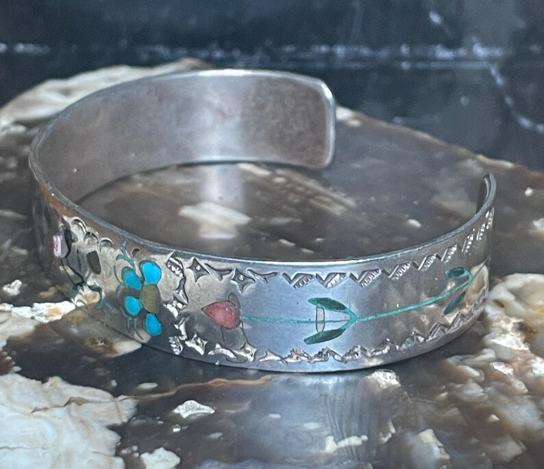May include: Silver cuff bracelet with etched floral and geometric designs. The bracelet features turquoise, red, and green accents. The open-ended design allows for easy wear. A unique piece of jewelry.