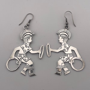 May include: A pair of silver-toned dangle earrings featuring a cutout design of a figure wearing a plumed helmet and holding a circular hoop in each hand. The figures have etched sunburst patterns on their clothing and fringe details on their legs.