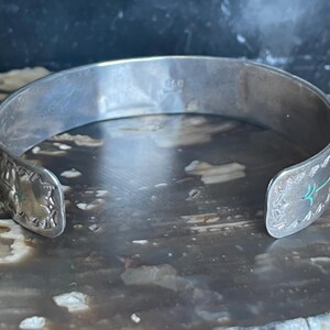 May include: A silver cuff bracelet with decorative edges and turquoise accents. The open-ended bracelet has a slightly curved shape and is designed to be worn on the wrist. The bracelet's surface has a polished, reflective quality.