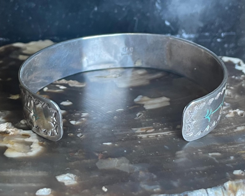 May include: A silver cuff bracelet with decorative edges and turquoise accents. The open-ended bracelet has a slightly curved shape and is designed to be worn on the wrist. The bracelet's surface has a polished, reflective quality.