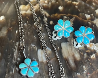 May include: A silver necklace with a flower pendant and matching flower earrings. The pendant and earrings feature a turquoise blue flower design. The jewelry is displayed on a brown and tan surface.