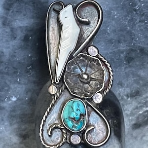 May include: A silver pendant featuring a bird-shaped mother-of-pearl inlay, a turquoise stone, and a floral design. The pendant has an ornate design with a rope-like border and small silver accents. The turquoise stone is oval-shaped.