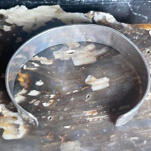 May include: A silver-toned cuff bracelet with a smooth, rounded design. The inside of the bracelet has a small marking. The bracelet is displayed on a surface with a marbled appearance.