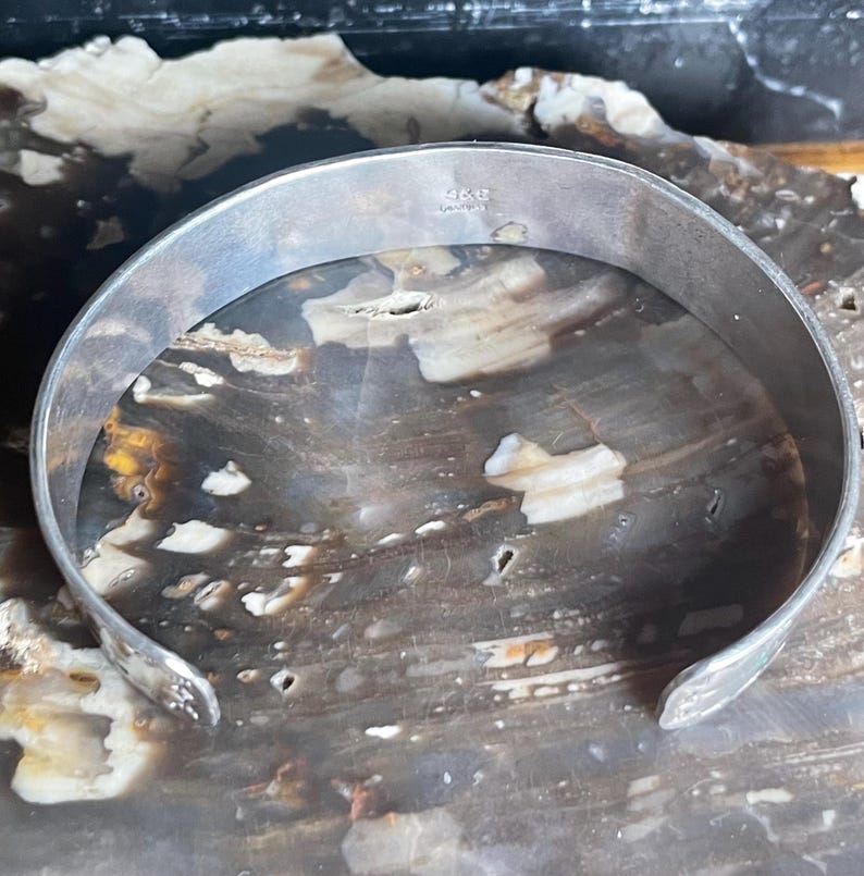 May include: A silver-toned cuff bracelet with a smooth, rounded design. The inside of the bracelet has a small marking. The bracelet is displayed on a surface with a marbled appearance.