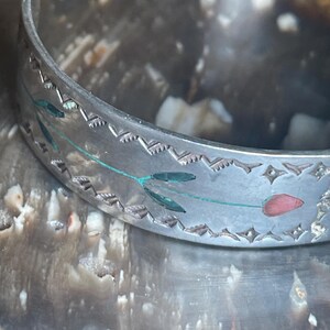 May include: Silver-toned cuff bracelet with etched floral and geometric designs. The bracelet features a delicate pattern of leaves and a red tulip. The edges are detailed with a repeating zigzag pattern.