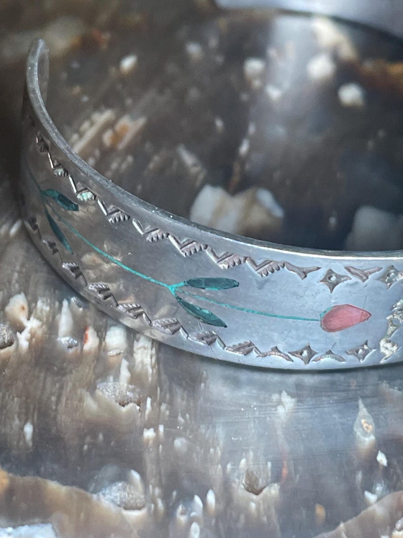 May include: Silver-toned cuff bracelet with etched floral and geometric designs. The bracelet features a delicate pattern of leaves and a red tulip. The edges are detailed with a repeating zigzag pattern.