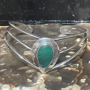 May include: A silver cuff bracelet with a central teardrop-shaped malachite stone. The bracelet has three parallel bands. The malachite stone is a vivid green colour, set in a silver bezel.