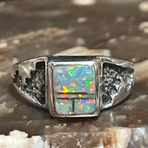 May include: A silver ring with a rectangular opal stone. The opal has a vibrant play of color, with flashes of green, blue, and yellow. The ring's band has a textured design on the sides.