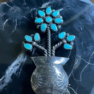 May include: A silver-toned metal flower pin with turquoise-colored stone accents. The pin features a vase-shaped base with floral designs and twisted stems supporting the turquoise flower blossoms. The pin is set against a dark, textured background.