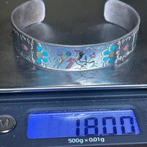 May include: Silver cuff bracelet with a bird and floral design. The bracelet features a detailed pattern with turquoise, pink, and blue accents. The bracelet is displayed on a digital scale.