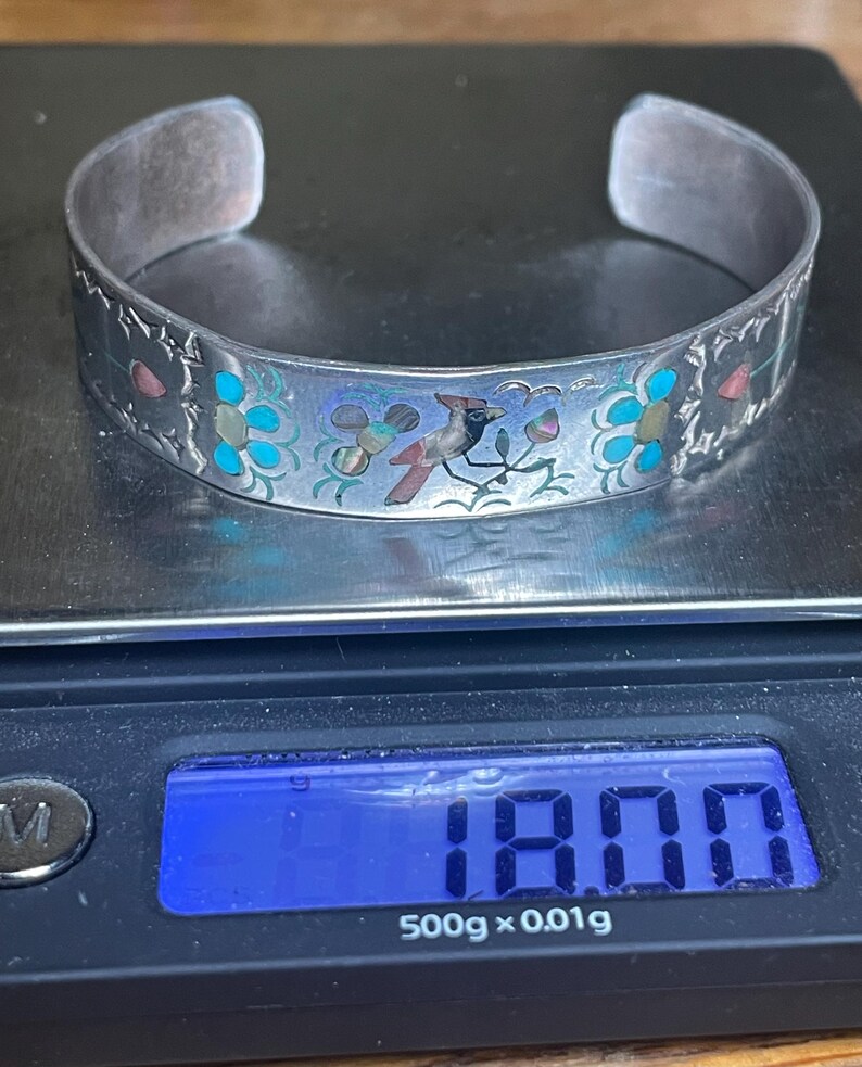 May include: Silver cuff bracelet with a bird and floral design. The bracelet features a detailed pattern with turquoise, pink, and blue accents. The bracelet is displayed on a digital scale.