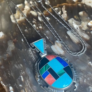 May include: A silver necklace with a pendant featuring a turquoise triangle and an oval design with turquoise, red, black, green, and blue inlays. The chain is a delicate silver link.