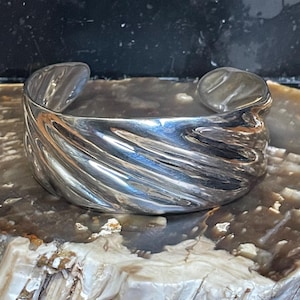 May include: A wide, silver-toned cuff bracelet with a textured, swirling design. The open-ended bracelet is displayed on a light-colored, textured surface. The cuff is approximately 2.5 inches wide.