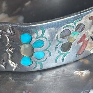 May include: Close-up of a silver bracelet with turquoise and abalone shell inlay. The bracelet features a floral design with turquoise petals and a central stone. The background is a blurred, textured surface.