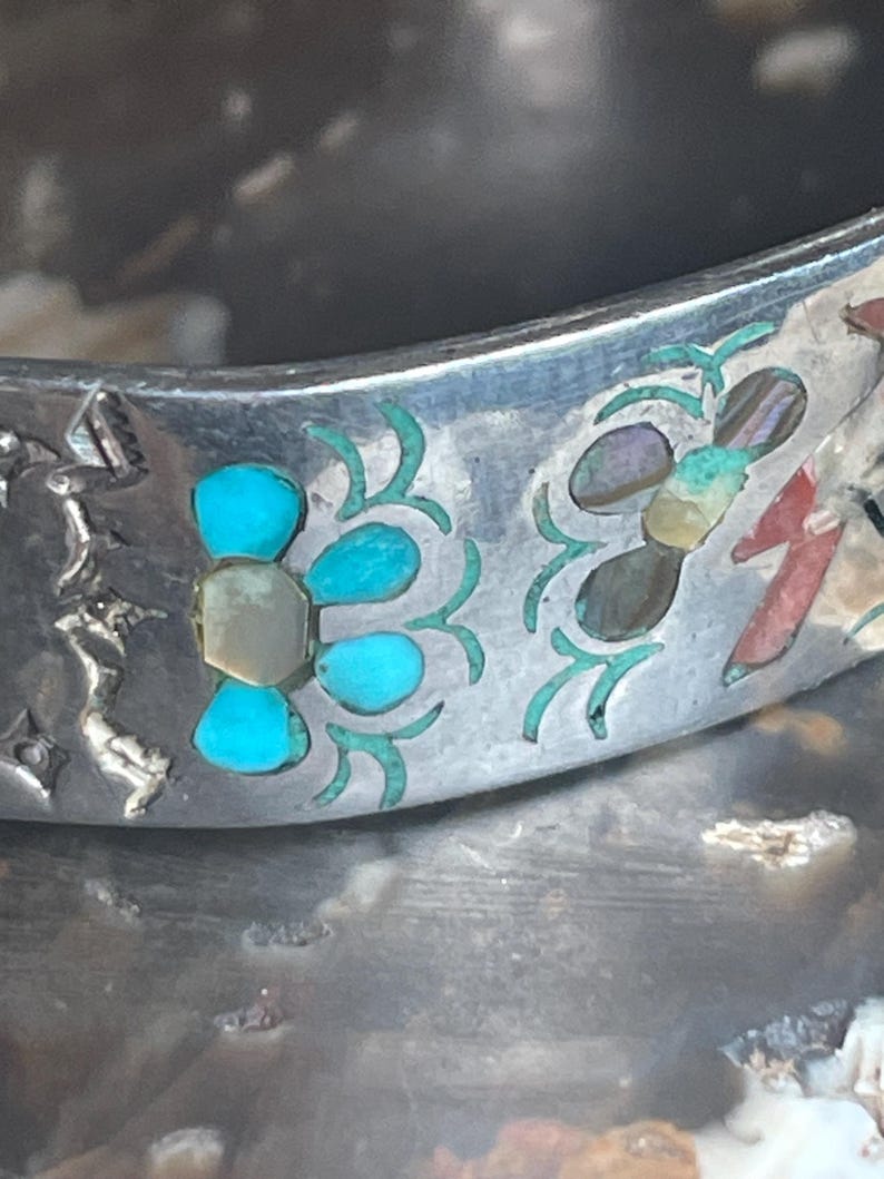 May include: Close-up of a silver bracelet with turquoise and abalone shell inlay. The bracelet features a floral design with turquoise petals and a central stone. The background is a blurred, textured surface.