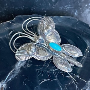 May include: A silver-toned butterfly brooch with a turquoise stone center. The brooch features detailed wing designs and a textured body. The butterfly is set against a dark, marbled background, highlighting its intricate craftsmanship.