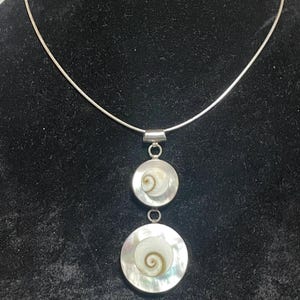 May include: A silver necklace featuring two circular pendants. Each pendant has a white and brown spiral design set within a silver frame. The necklace hangs on a delicate silver chain, creating an elegant accessory.