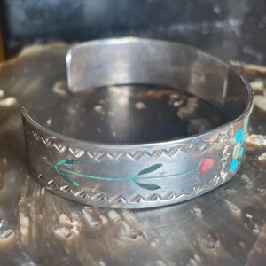 May include: Silver cuff bracelet with etched floral and leaf designs. The bracelet features inlaid turquoise, red, and green accents. The open design allows for easy wear. A unique piece of jewelry.
