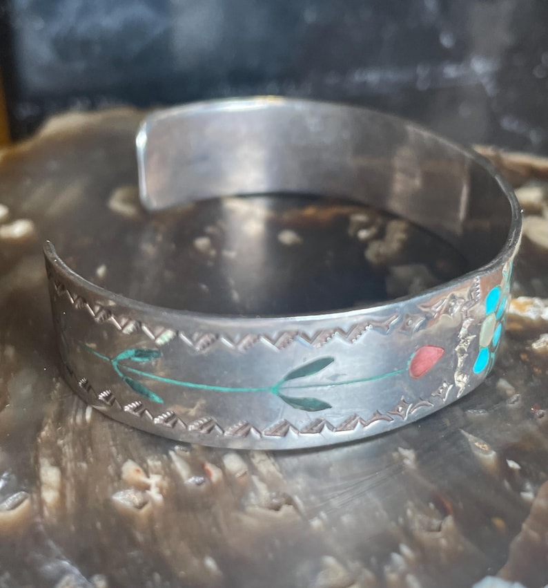 May include: Silver cuff bracelet with etched floral and leaf designs. The bracelet features inlaid turquoise, red, and green accents. The open design allows for easy wear. A unique piece of jewelry.