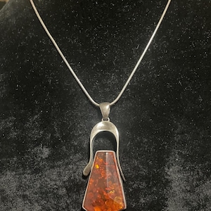 May include: A silver pendant necklace featuring a large, amber-colored stone. The stone is encased in a silver frame with a unique, elongated design. The necklace hangs on a delicate silver chain, creating a striking contrast between the warm amber and cool silver.