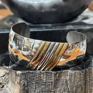 Puede incluir: Una pulsera de plata con un diseño diagonal dorado y plateado. La pulsera es ancha y tiene un acabado pulido. Las secciones doradas tienen una textura cepillada. Joyería.