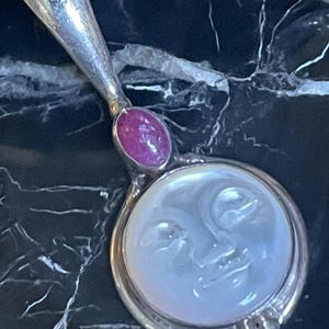 May include: A silver-toned hand mirror with a handle and a round face. The face of the mirror has a carved moon design. A pink gemstone is set in the handle. The mirror is on a black marble surface.