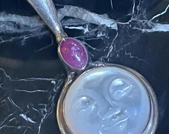 May include: A silver-toned hand mirror with a handle and a round face. The face of the mirror has a carved moon design. A pink gemstone is set in the handle. The mirror is on a black marble surface.