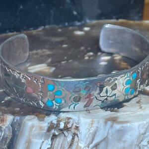 May include: Silver cuff bracelet with intricate floral and bird designs. The bracelet features turquoise, red, and green accents. The open design allows for easy wear. A unique piece of jewelry.