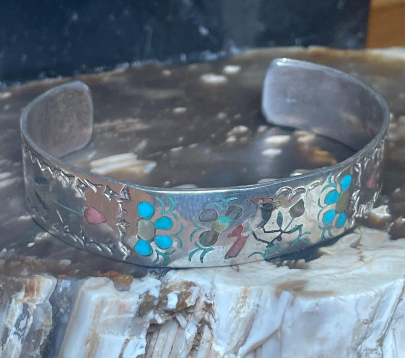 May include: Silver cuff bracelet with intricate floral and bird designs. The bracelet features turquoise, red, and green accents. The open design allows for easy wear. A unique piece of jewelry.