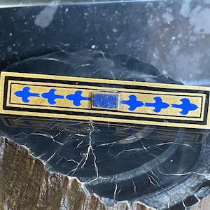 May include: A rectangular gold-toned bar pin with a black border and blue fleur-de-lis designs. The pin features a rectangular blue stone in the center and is set against a dark, textured surface.