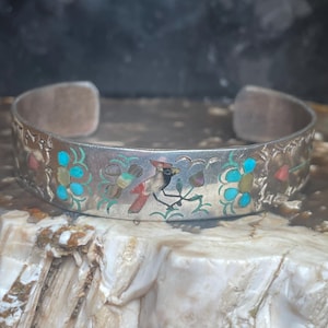 Op de afbeelding: Zilveren manchetarmband met ingelegde vogel- en bloemmotieven. De armband heeft een kardinaal met rode, zwarte en witte veren, omringd door turquoise en koraal accenten. De randen hebben een decoratief patroon.