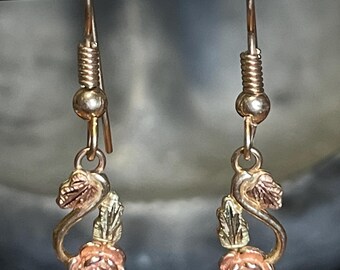 May include: A pair of gold-toned earrings featuring a rose design. Each earring has a small rose with pink petals and green leaves. The earrings are suspended from a hook with a coiled detail.