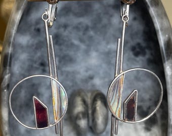 May include: A pair of silver-toned clip-on earrings. Each earring features a circular design with iridescent abalone shell and a burgundy-colored geometric shape. Vertical bars and a long, thin drop complete the design.