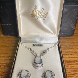 May include: A vintage jewelry set by Lang, featuring a necklace and matching earrings. The set includes a cameo pendant and earrings, all set in sterling silver. The jewelry is presented in a satin-lined box with the Lang logo.