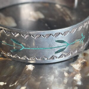 May include: A silver-toned cuff bracelet with a decorative border and turquoise-colored leaf accents. The bracelet has a detailed, engraved design and a slightly worn appearance, suggesting it is vintage or handmade.