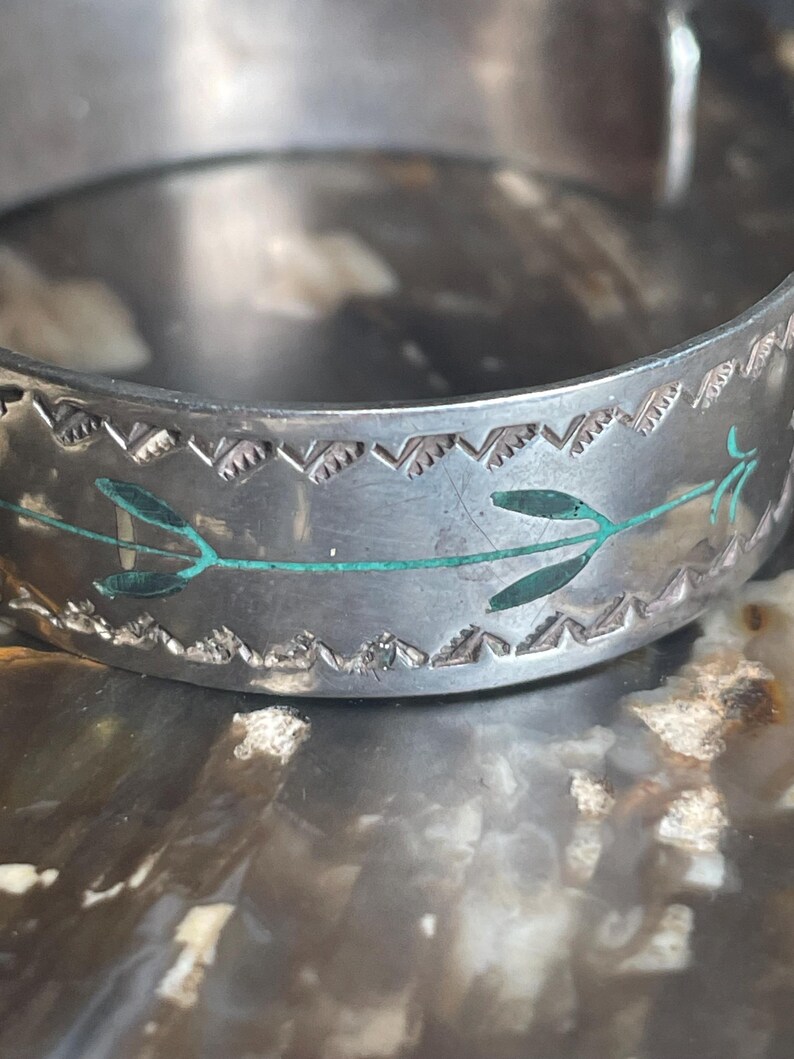 May include: A silver-toned cuff bracelet with a decorative border and turquoise-colored leaf accents. The bracelet has a detailed, engraved design and a slightly worn appearance, suggesting it is vintage or handmade.